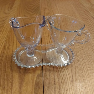 May include: A clear glass creamer and sugar set with beaded edges, sitting on a matching beaded tray.