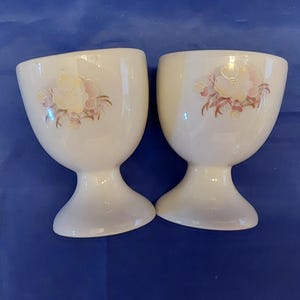 May include: Two white ceramic egg cups with a floral design in pink and yellow. The egg cups have a rounded top and a flared base. The background is a solid blue.