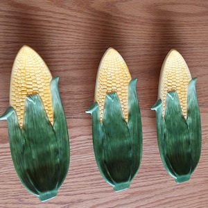 May include: Three ceramic corn-shaped dishes featuring yellow kernels and textured green husks. Each dish is elongated and molded to resemble a fresh ear of corn, with detailed husk leaves wrapping around the base and sides of the yellow cob section.