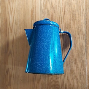 May include: A blue enamelware coffee pot with a lid and handle. The pot has a speckled finish and a spout for pouring. The handle is curved and attached to the side of the pot. The pot is approximately 10 inches tall.