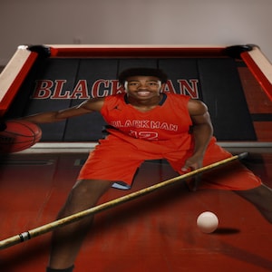 May include: A basketball player wearing an orange jersey with the text "Blackman" on the front, dribbling a basketball on a pool table with a red felt surface. The player is holding a pool cue in their right hand.