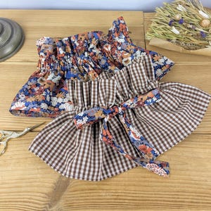 Puede incluir: Dos piezas de tela con volantes. Una presenta un estampado floral en azul, naranja y blanco. La otra es de cuadros marrones y blancos con un lazo floral. Ambas est&aacute;n sobre una superficie de madera.