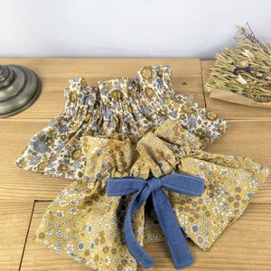 May include: Two floral fabric cuffs with elasticated tops and a blue bow. One cuff is mustard yellow with white and blue flowers, the other is a similar pattern in blue, brown, and white. The cuffs are on a wooden surface.