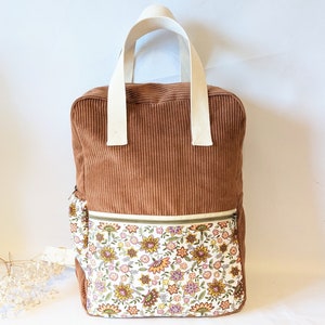 Peut inclure: Sac à dos en velours côtelé marron avec une poche à imprimé floral et des poignées crème. Le sac à dos présente une poche avant zippée avec un motif floral coloré dans les tons de jaune, orange et violet. Le sac est de forme rectangulaire.