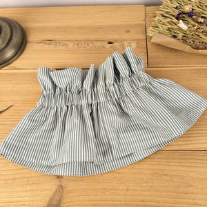 May include: A gathered, striped fabric skirt in shades of gray and white. The skirt has a ruffled top and a flared bottom. The fabric is lightweight and appears to be cotton or a similar material. The skirt is displayed on a wooden surface.