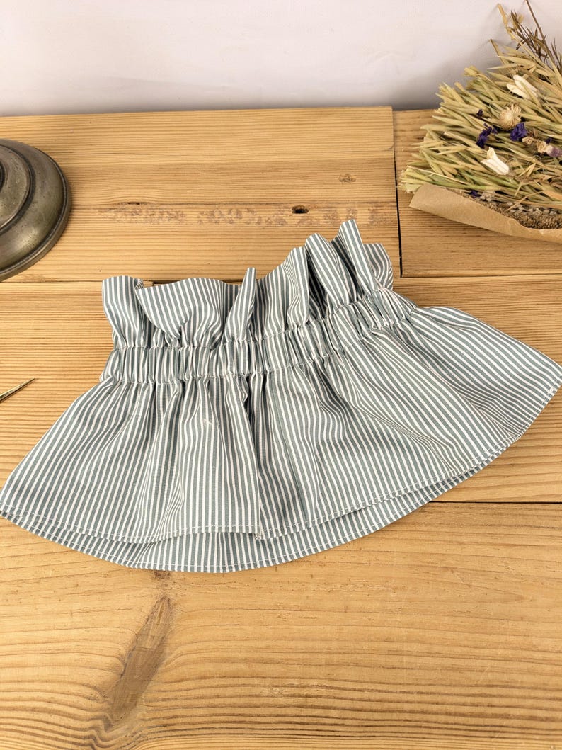 May include: A gathered, striped fabric skirt in shades of gray and white. The skirt has a ruffled top and a flared bottom. The fabric is lightweight and appears to be cotton or a similar material. The skirt is displayed on a wooden surface.