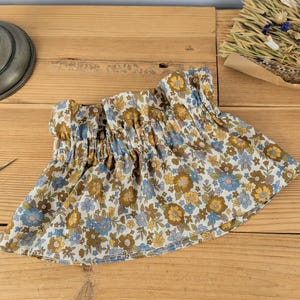 May include: A floral print skirt with a gathered waistband. The skirt features a pattern of blue, yellow, and brown flowers on a cream background. The fabric is gathered at the waist, creating a ruffled effect. The skirt is displayed on a wooden surface.