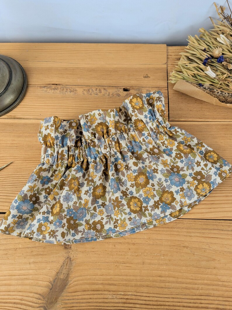 May include: A floral print skirt with a gathered waistband. The skirt features a pattern of blue, yellow, and brown flowers on a cream background. The fabric is gathered at the waist, creating a ruffled effect. The skirt is displayed on a wooden surface.