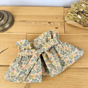 May include: A pair of beige fabric wrist cuffs with a floral pattern in pink, blue, and yellow. The cuffs are tied together with a matching fabric tie, and are displayed on a wooden surface.