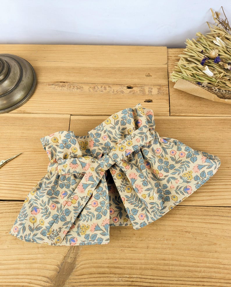 May include: A pair of beige fabric wrist cuffs with a floral pattern in pink, blue, and yellow. The cuffs are tied together with a matching fabric tie, and are displayed on a wooden surface.