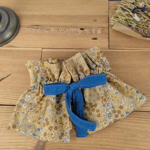 May include: A yellow floral fabric garment with a blue bow. The fabric features small white and blue flowers. The bow is made of solid blue fabric. The garment is ruffled at the top and bottom, with a vintage aesthetic.