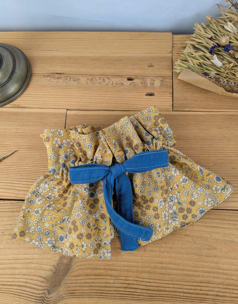 May include: A yellow floral fabric garment with a blue bow. The fabric features small white and blue flowers. The bow is made of solid blue fabric. The garment is ruffled at the top and bottom, with a vintage aesthetic.