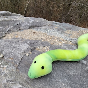 May include: A green and yellow plush snake toy with a smiling face and black eyes. The snake is lying on a gray rock.