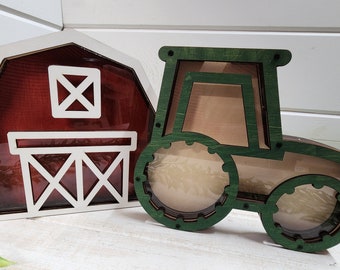Op de afbeelding: Twee houten spaarpotten, een in de vorm van een rode schuur en de andere in de vorm van een groene tractor. Beide hebben heldere kunststof ramen om de munten erin te zien.