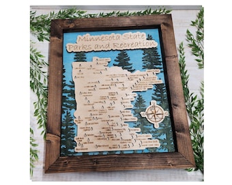 Op de afbeelding: Een houten kaart van Minnesota State Parks and Recreation, met een kompas in de rechterbenedenhoek. De kaart is ingelijst in een donkerhouten lijst en heeft een blauwe achtergrond met groene bomen.