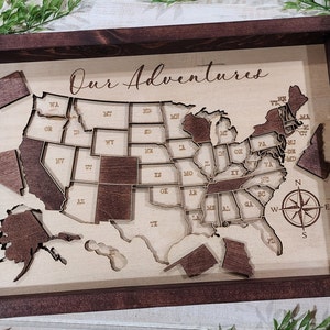 May include: Wooden United States map with state outlines and removable pieces, framed in dark brown. The words "Our Adventures" are scripted across the top. A compass rose is etched in the lower right corner. States are labeled with their abbreviations.
