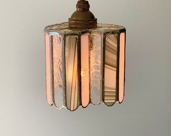 Op de afbeelding: Een hanglamp met een heldere glazen kap met een gestreept patroon in roze en witte tinten. Het licht is aan en verlicht de kamer.