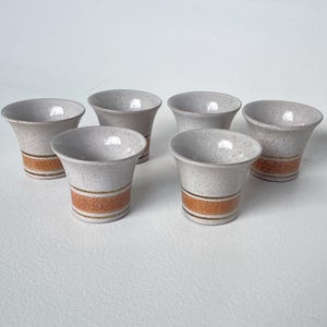 May include: A set of six ceramic cups with a speckled off-white finish. Each cup features a flared rim and a wide, textured orange band around the lower section, accented by thin dark brown horizontal lines that encircle the base of the vessel.