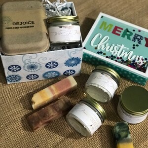 May include: A gift box filled with bath and body products. The box is decorated with snowflakes and has a brown lid with the text "REJOICE COCONUT & KINGS-PEPPERMINT BARK". The box contains three jars of bath products and three bars of soap. The jars are labeled "White Tea & Pear", "This is the Place", and "Salt Dream". The soap bars are in various colors and patterns. The box is sitting on a burlap surface next to a sign that says "Merry Christmas" in colorful letters.