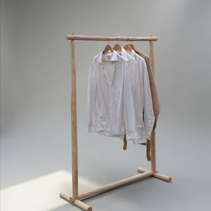 May include: A light-coloured wooden clothing rack with a simple frame construction. Hanging on the rack are two white long-sleeved button-down shirts and a tan-coloured textured garment, each suspended on a wooden hanger from the top horizontal bar.