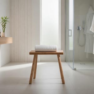 Puede incluir: Interior de baño minimalista con un taburete de madera y una toalla blanca enrollada. La habitación tiene paredes blancas, una ducha de cristal y un tocador de madera. Dos albornoces blancos cuelgan de la pared.