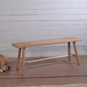 May include: A light-colored wooden bench with four legs and a connecting support beam. The bench is set against a white-painted wood plank wall. A yellow Labrador Retriever is lying on the floor in the foreground.