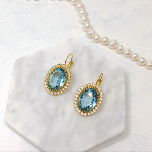 May include: A pair of gold earrings with a large oval-shaped blue gemstone surrounded by smaller clear gemstones.