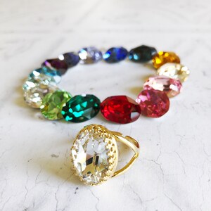 May include: A gold ring with a large clear crystal stone set in a gold bezel. The ring is surrounded by a circle of smaller crystals in a rainbow of colors.