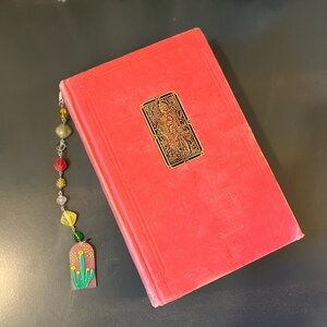 May include: A red hardcover book with a gold bookmark featuring a detailed illustration. The bookmark is attached to a beaded chain with colorful beads and a charm of a green cactus with yellow flowers.