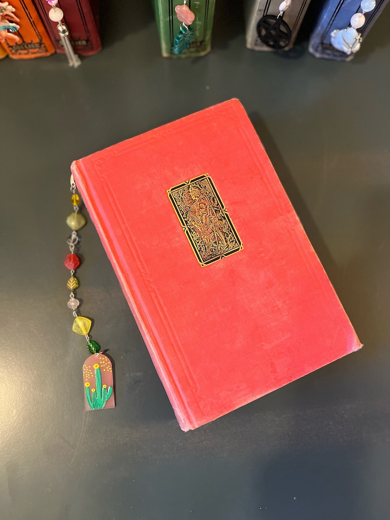 May include: A red hardcover book with a gold bookmark featuring a detailed illustration. The bookmark is attached to a beaded chain with colorful beads and a charm of a green cactus with yellow flowers.