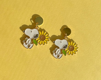 May include: A pair of Snoopy earrings. Each earring features a white Snoopy figure holding a yellow sunflower with green leaves. The earrings have gold-colored posts and are set against a yellow background.