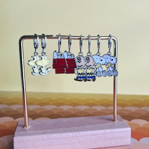 May include: A display stand holding six pairs of cartoon character earrings. The earrings feature Snoopy, Woodstock, and other Peanuts characters in various poses. The stand has a light pink wooden base and a gold-colored metal frame.
