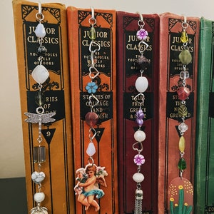 May include: Five bookends with different charms and beads hanging from them. The charms include a dragonfly, a cherub, a flower, a heart, and a cactus. The beads are made of glass, metal, and wood. The bookends are all made of metal and have a silver finish.