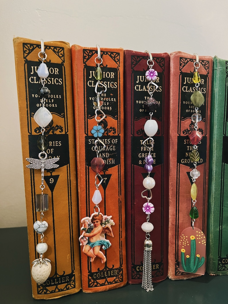 May include: Five bookends with different charms and beads hanging from them. The charms include a dragonfly, a cherub, a flower, a heart, and a cactus. The beads are made of glass, metal, and wood. The bookends are all made of metal and have a silver finish.
