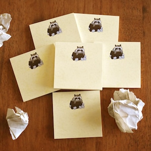 Puede incluir: Seis notas adhesivas beige con una ilustración de mapache en cada una. El mapache está sentado en una caja con una flor blanca.