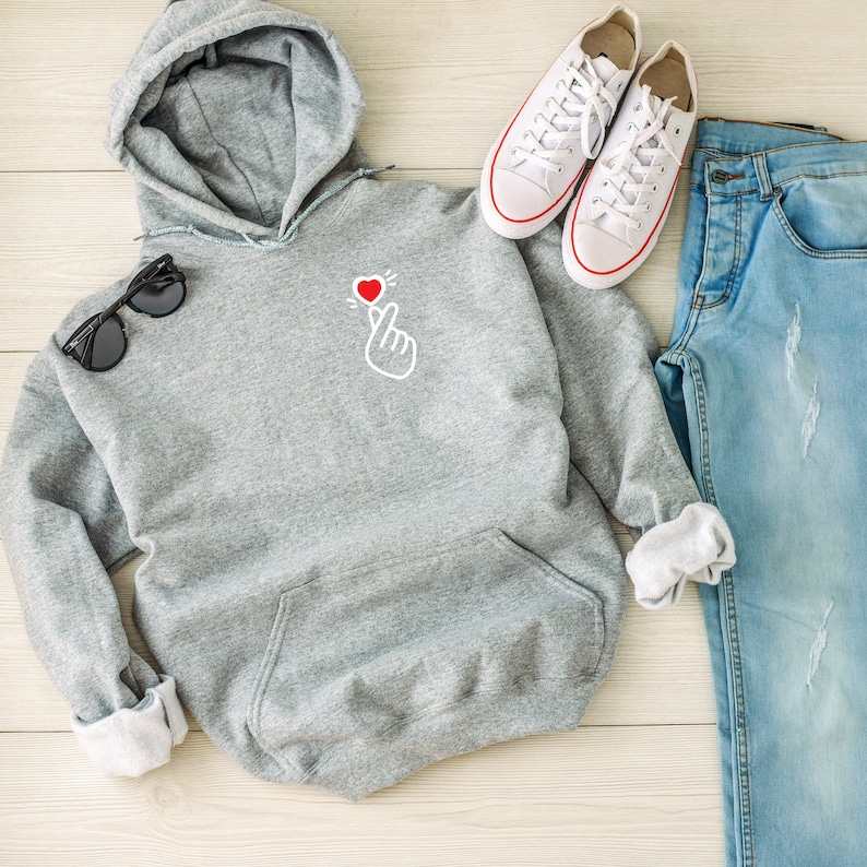 May include: A gray hoodie with a small red heart and a hand gesture on the front. The hoodie is laying on a white wooden surface with a pair of white sneakers and blue jeans.
