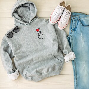 May include: A gray hooded sweatshirt with a small red heart and a hand gesture on the front. The sweatshirt is laid out on a wooden surface with a pair of white sneakers and blue jeans.