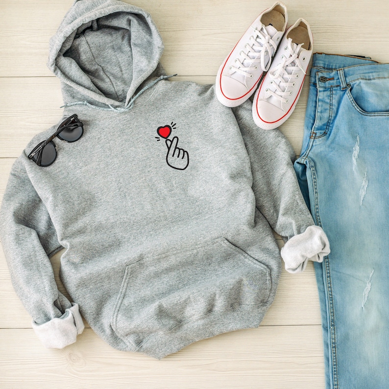 May include: A gray hooded sweatshirt with a small red heart and a hand gesture on the front. The sweatshirt is laid out on a wooden surface with a pair of white sneakers and blue jeans.