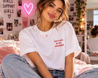 May include: A white short-sleeved t-shirt featuring the red text "too cute to be straight" printed on the left chest area. The shirt has a crew neckline and is worn with blue denim jeans. The fabric appears to be a standard cotton blend.