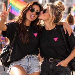 Pode incluir: Duas camisetas pretas com um pequeno triângulo rosa sólido no peito esquerdo. Cada peça tem gola redonda e mangas curtas, usadas com shorts jeans e mochilas pretas em um ambiente externo iluminado.