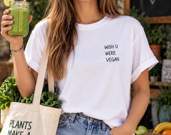 May include: A white t-shirt with the black text WISH U WERE VEGAN printed on the chest. A beige canvas tote bag features the black text PLANTS MAKE A HAPPIER PLANET with a heart symbol. The shirt has short sleeves and is worn with blue jeans.