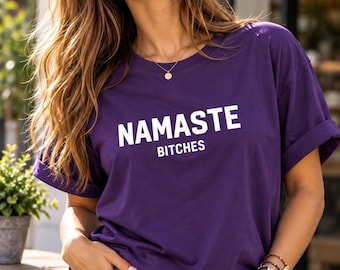 May include: A purple short-sleeved t-shirt featuring the white text NAMASTE BITCHES printed across the chest. The shirt has a crew neckline and rolled sleeves, paired with light blue distressed denim jeans with visible rips.