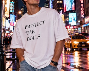 Puede incluir: Una camiseta blanca de manga corta con el texto negro "PROTECT THE DOLLS" impreso en el pecho. La prenda cuenta con cuello redondo y un corte holgado, combinada con pantalones vaqueros azul claro y un reloj de pulsera negro.