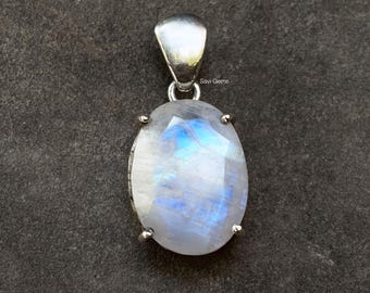May include: Oval-shaped moonstone pendant with a silver bail and setting. The moonstone has a blue and white iridescent glow. The pendant is set against a dark gray background. The text "Savi Gems" is visible.