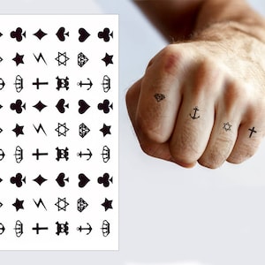 May include: A sheet of temporary tattoos with a variety of small black designs, including hearts, diamonds, stars, anchors, crosses, and other symbols. The tattoos are shown applied to a person's fingers.