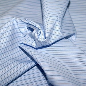 May include: Close-up of light blue fabric with thin, horizontal stripes in varying shades of blue and white. The fabric is gathered and folded, creating shadows and texture. The material appears to be cotton or a similar lightweight textile.