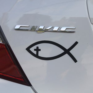 Puede incluir: Un maletero de coche blanco con un emblema "CIVIC" cromado y una pegatina negra con el símbolo Ichthys. La pegatina presenta un contorno de pez con una cruz en su interior. El piloto trasero rojo del coche es visible a la izquierda.