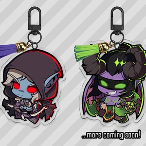 Op de afbeelding: Twee sleutelhangers met een paarse en groene kwast. De sleutelhanger links toont een elf met blauwe huid en rode ogen die een zwarte kapmantel draagt. De sleutelhanger rechts toont een demon met paarse huid, groene ogen en hoorns die een groen-zwarte outfit draagt. De tekst "...more coming soon!" staat onderaan de afbeelding.