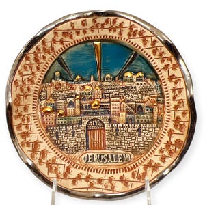 Puede incluir: Un plato decorativo redondo de cerámica con una representación en relieve de la ciudad de Jerusalén. El centro muestra un paisaje urbano detallado con cúpulas pintadas en oro sobre un cielo azul, enmarcado por un borde texturizado con la palabra JERUSALEM.