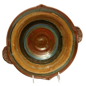 Könnte beinhalten: Eine große, runde Keramikschüssel mit zwei Griffen. Die Schüssel ist mit brauner, orangefarbener und blauer Glasur verziert, die ein wirbelndes Muster erzeugt. Die Schüssel ist perfekt zum Servieren von Salaten, Pasta oder anderen Gerichten.